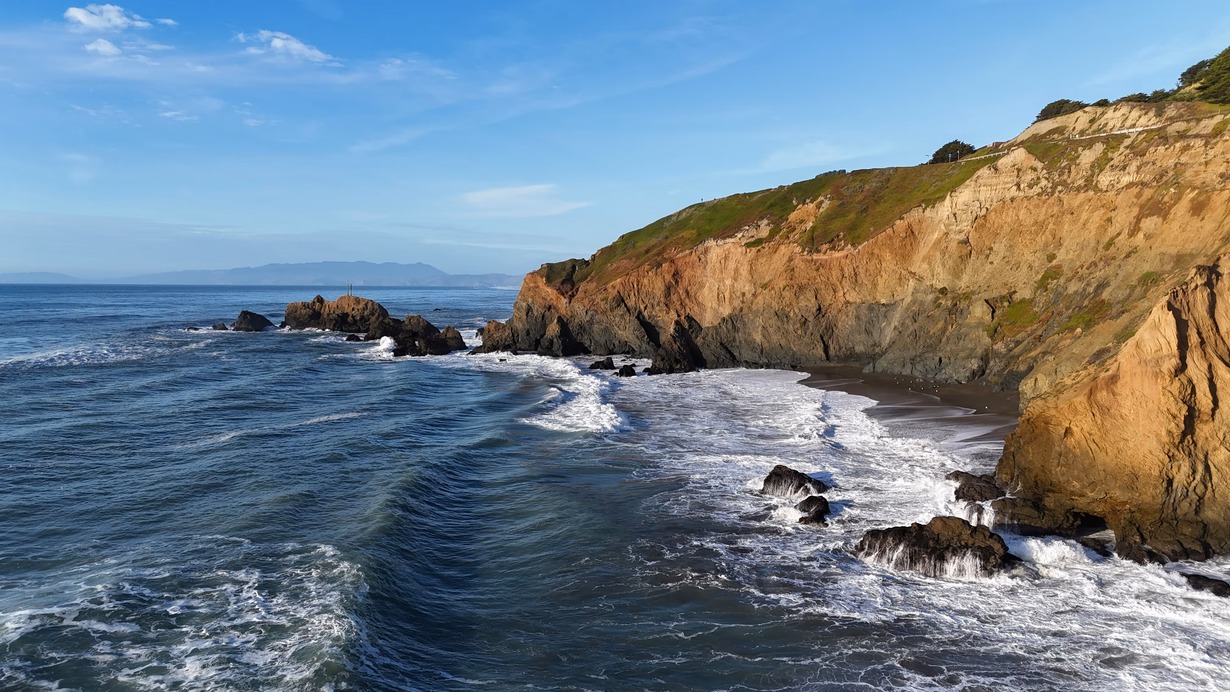 Ocean waves crashing against rocky cliffs with a small sandy beach under a clear blue sky.