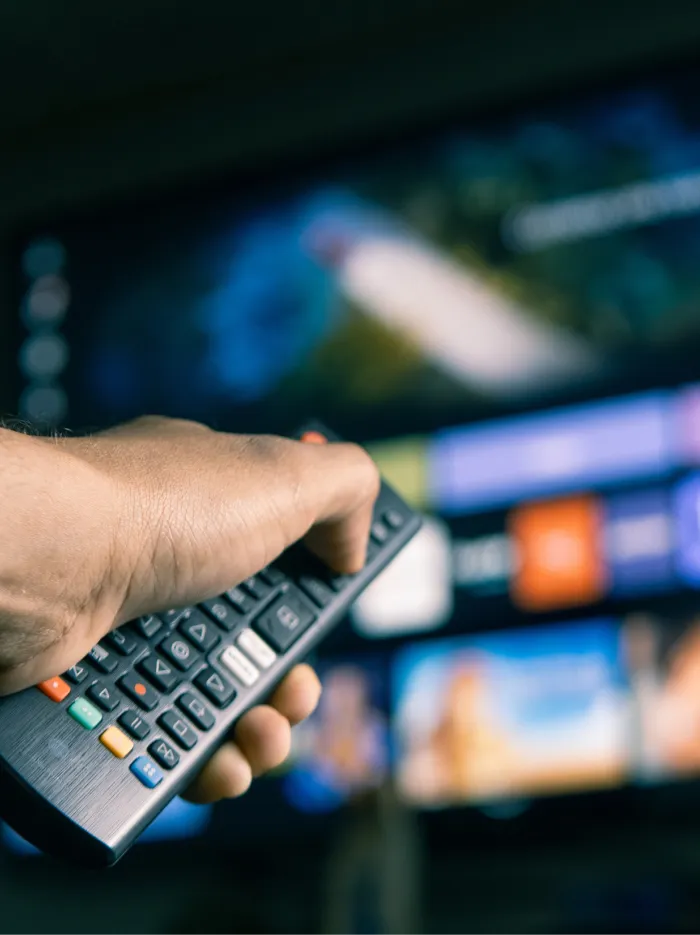 Hand holding a TV remote control pointed at a large screen with various colorful blurred app icons.