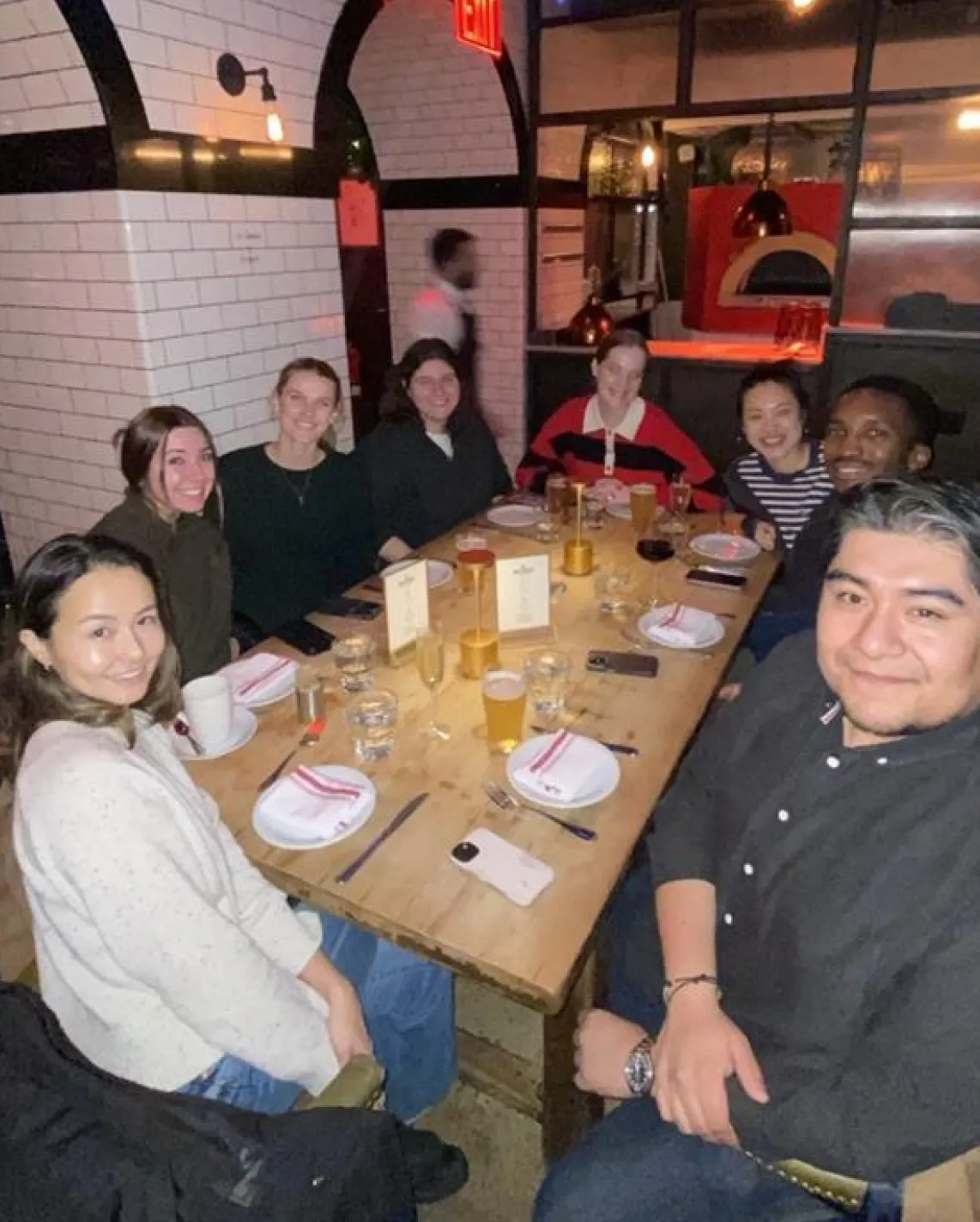 Group of eight people smiling and seated around a wooden restaurant table with drinks and menus.