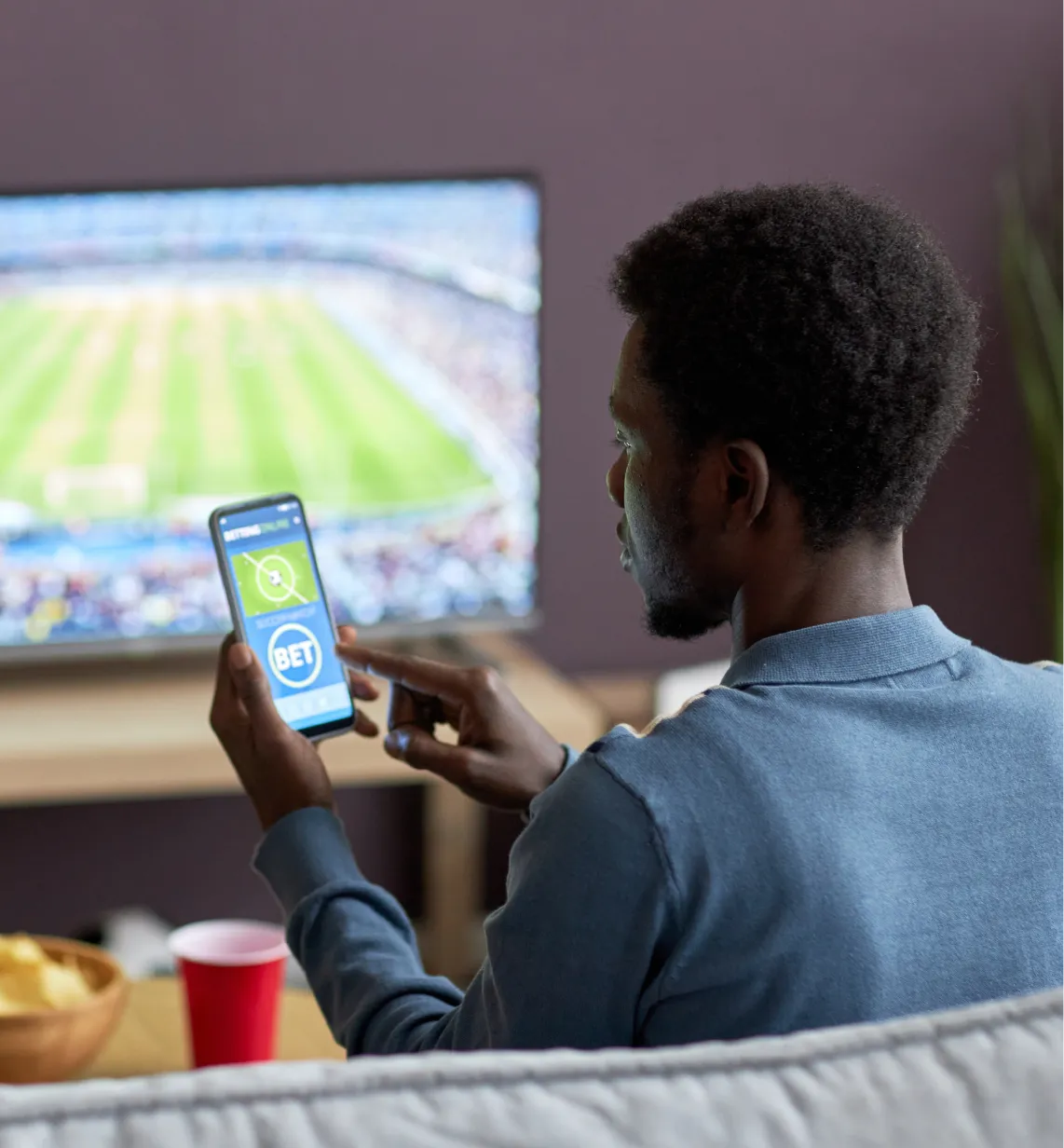 Man using a sports betting app on his smartphone while watching a football match on TV.