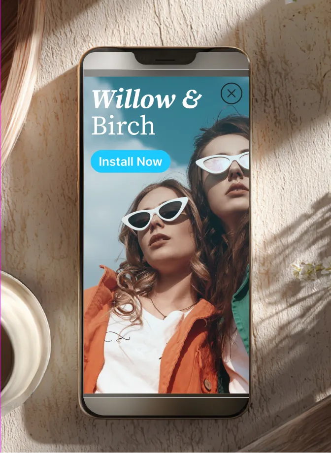Smartphone on a table displaying an ad with two women wearing spring jackets and sunglasses for Willow & Birch and an Install Now button.
