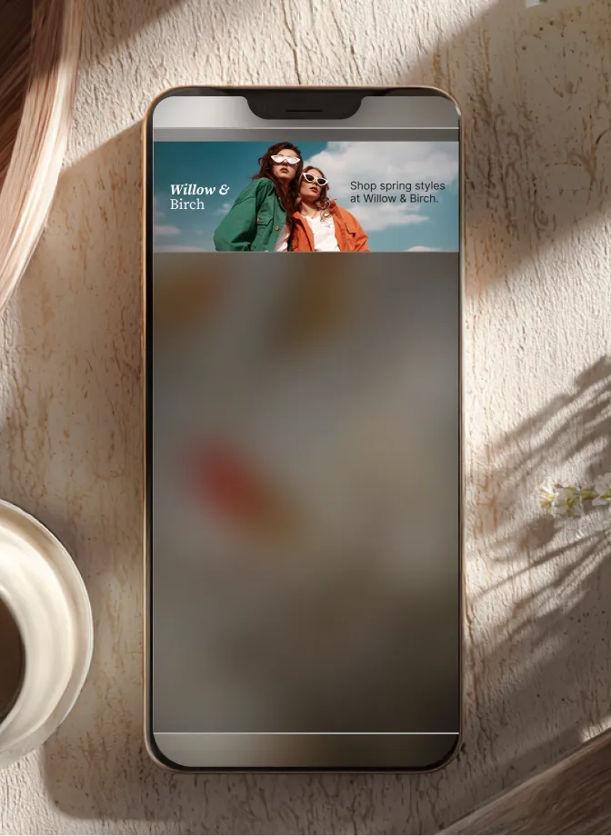 Smartphone on textured surface displaying Willow & Birch ad with two women in sunglasses modeling spring clothes.