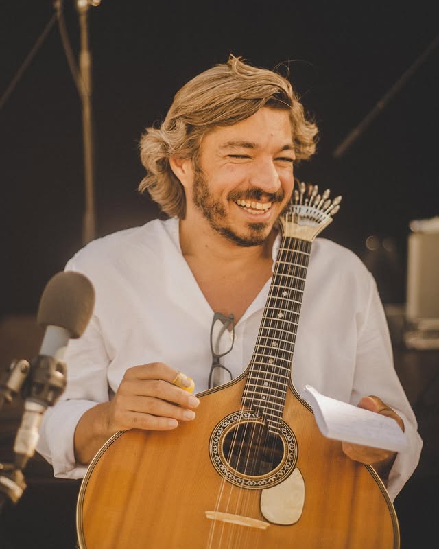 A men in a white t-shirt holding a Portuguese guitar with a microphone in front of him