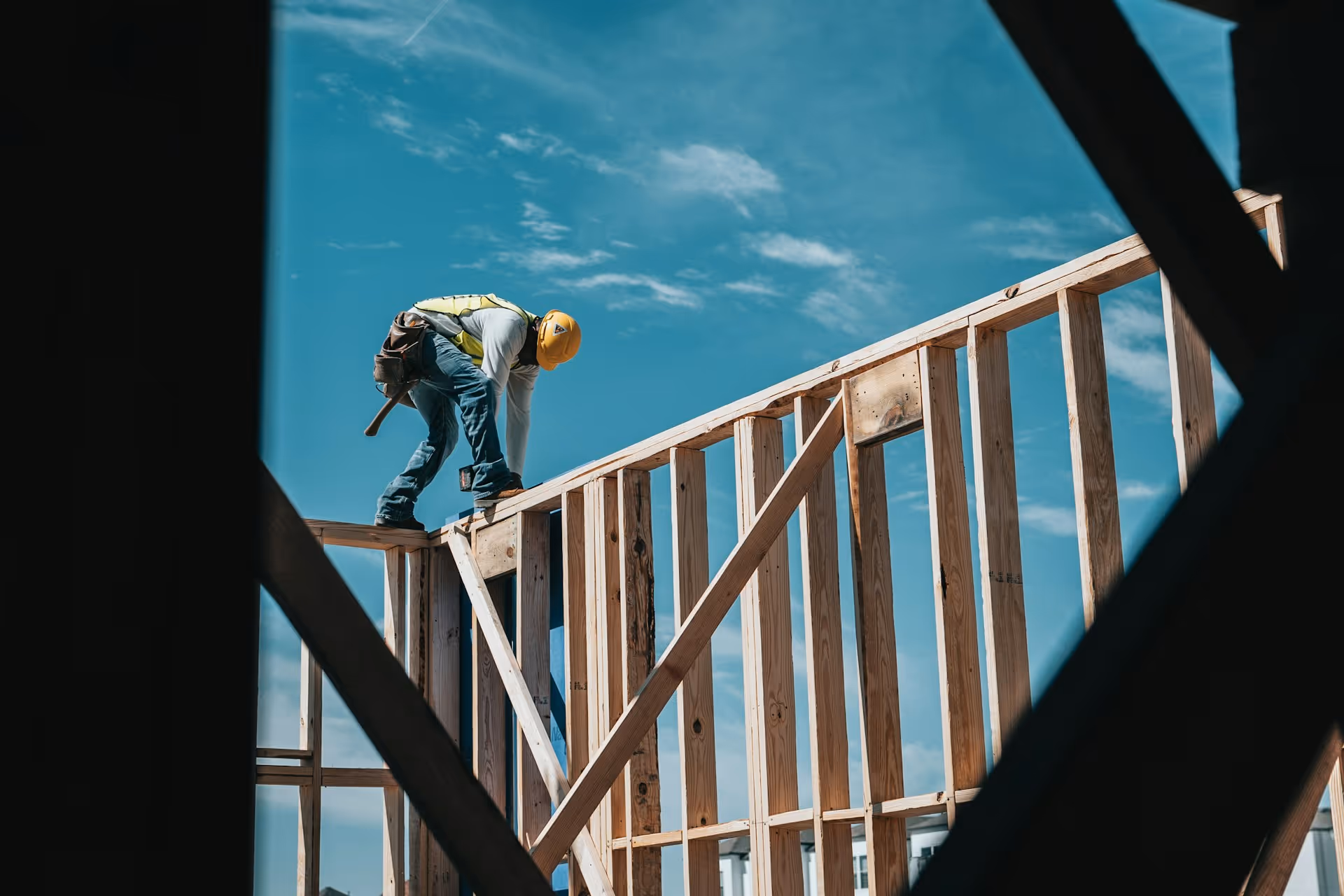 Construction worker wearing a yellow hard hat and safety vest bending over a wooden frame structure under a blue sky.