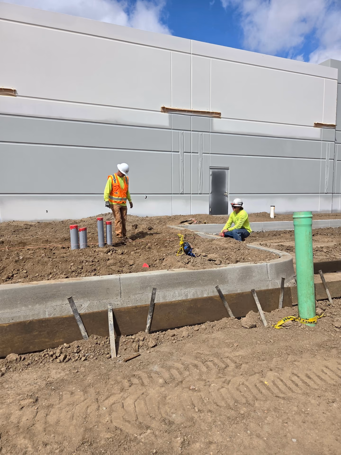 Two construction workers wearing safety helmets and bright yellow-green shirts at a construction site with dirt and concrete curbs near a large gray building under a partly cloudy sky. One worker is standing near capped pipes, and the other is sitting on a curb section. A tall green pipe is visible in the foreground with yellow caution tape.
