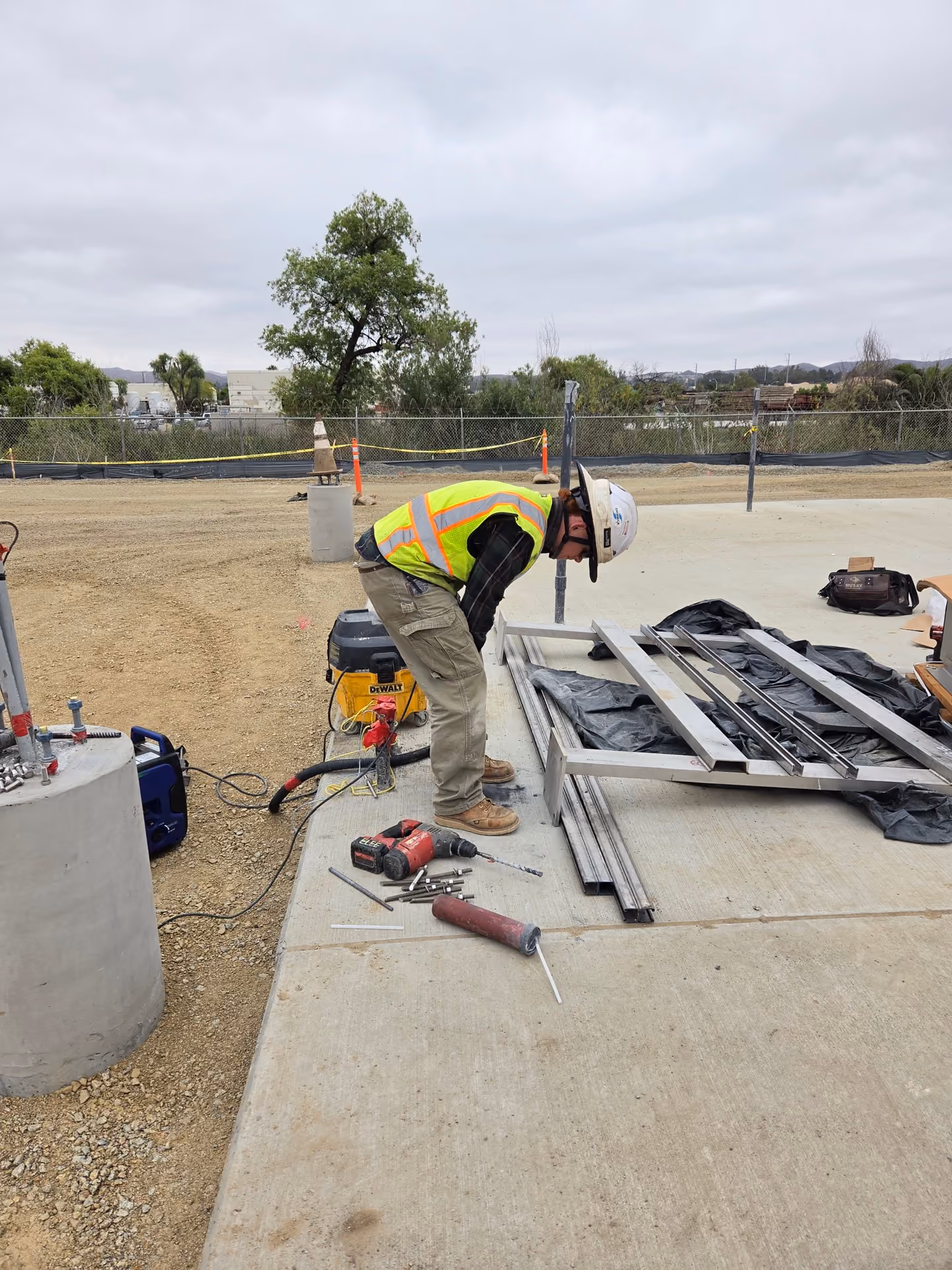 Construction worker wearing a white hard hat, yellow safety vest, and tan pants bending over on a concrete surface at a construction site with tools and metal frames around him.