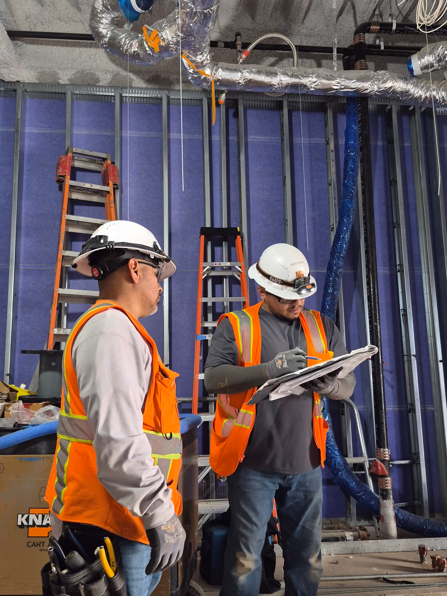 Two construction workers wearing white hard hats, orange safety vests, and gloves inside a building under construction; one worker is writing on a clipboard while the other looks on, with ladders, exposed wall framing, and insulated ductwork visible in the background.