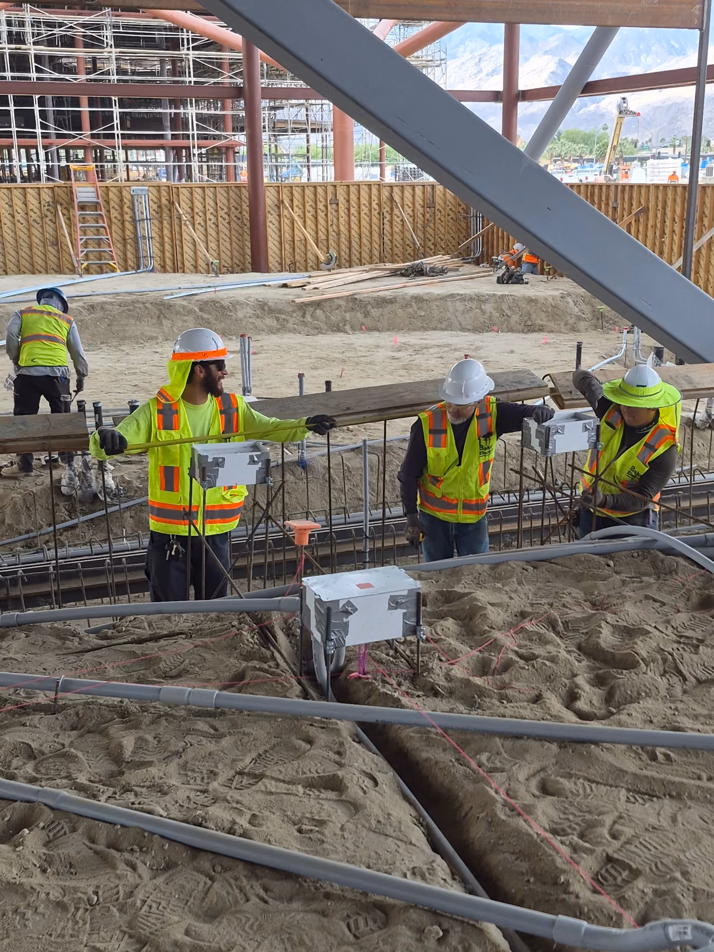 Three construction workers wearing safety vests and helmets stand on a sandy construction site with exposed wiring and pipes, holding a long wooden beam under a metal framework with mountains visible in the background.