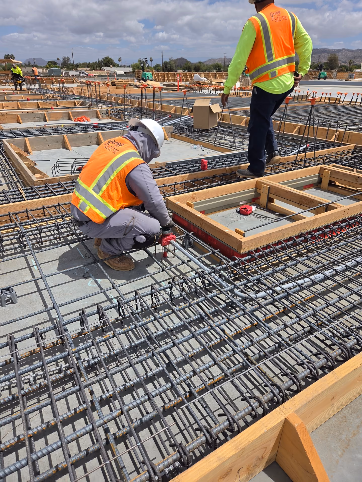 Two construction workers wearing orange safety vests labeled Gregg Electric work on a concrete foundation with steel rebar and electrical conduits, surrounded by wooden formwork under a partly cloudy sky.