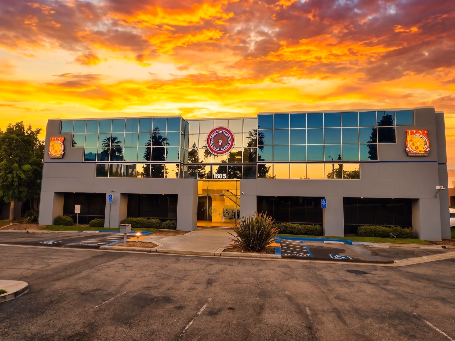 Modern two-story office building with reflective windows at sunset, showing palm tree reflections and three circular signs above the entrance labeled 1605; parking spaces and handicap signs are visible in front.