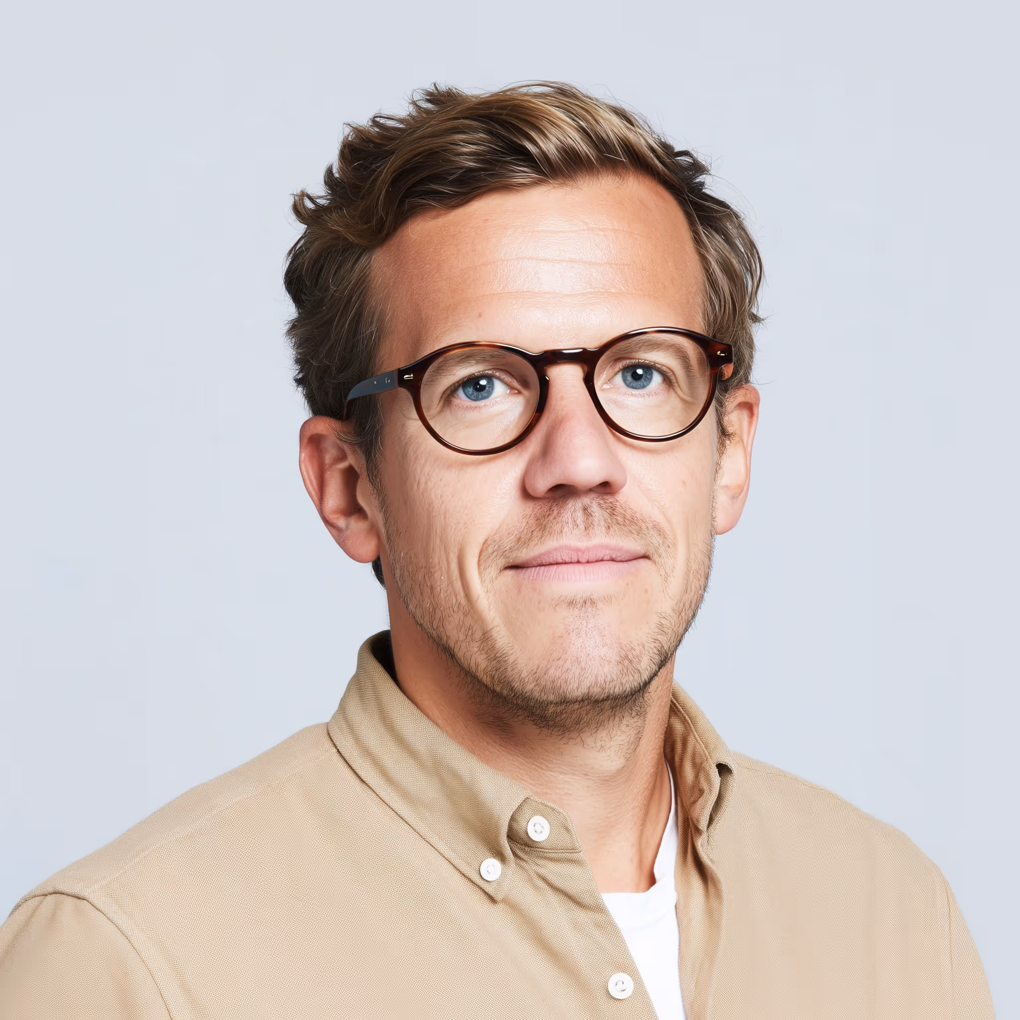 Man with light brown hair, blue eyes, and glasses wearing a beige button-up shirt over a white t-shirt.
