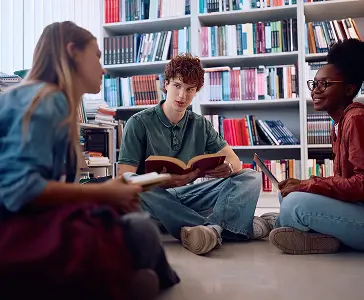 Three students sitting on floor discussing books in library