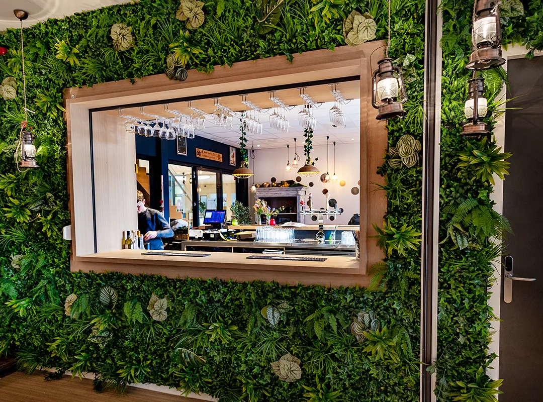 Groene kunstplantenwand rondom een horecakiosk met doorkijkvenster en sfeervolle lantaarns