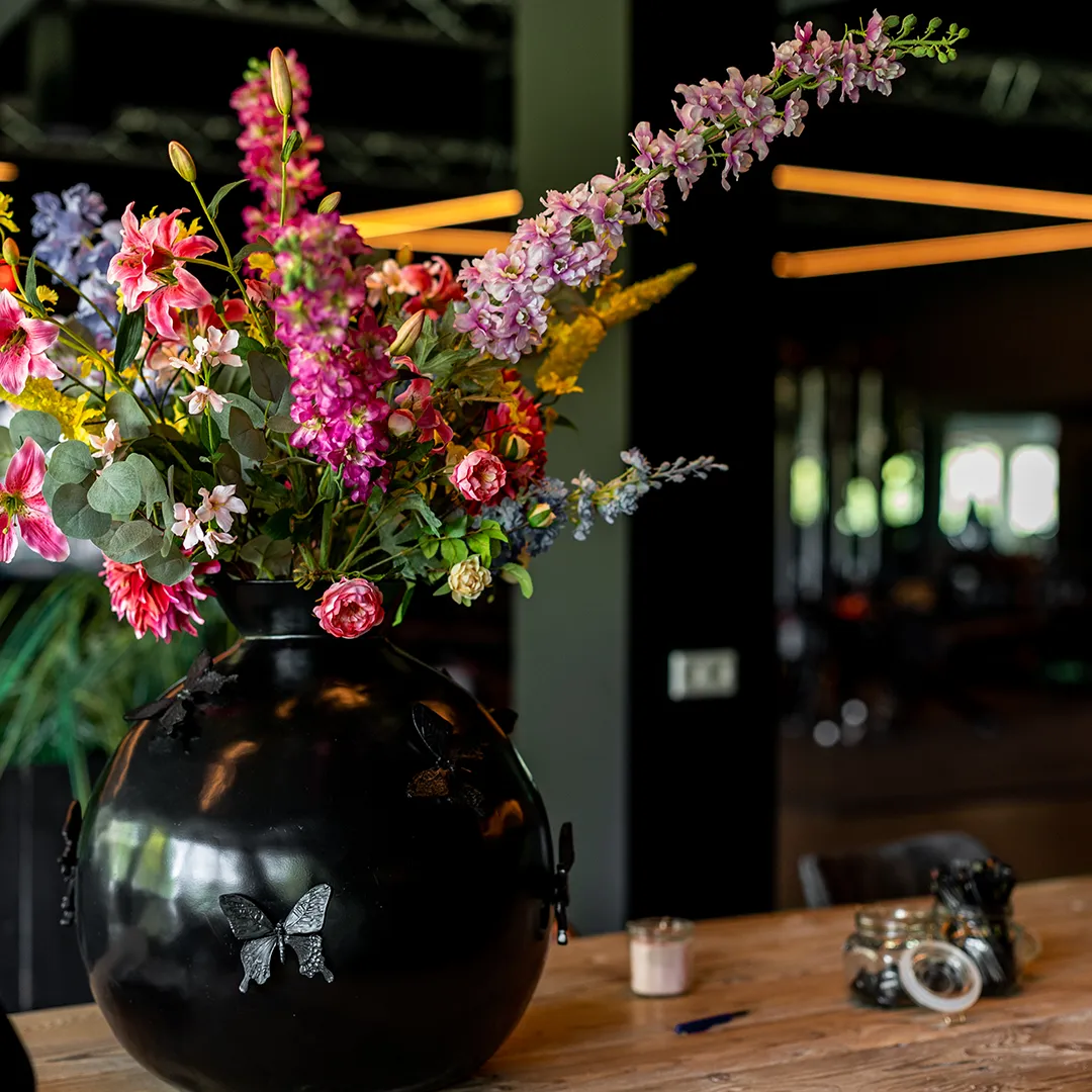 Kleurrijk bloemstuk in zwarte vaas als tafelstuk in een donker horecainterieur, van Miverde