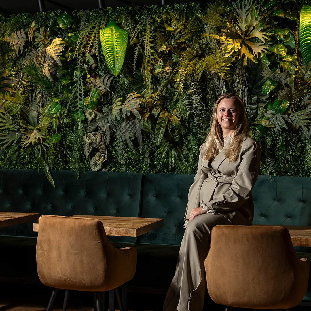 Blonde vrouw in beige pak zittend aan tafel met verticale plantenwand en groene bank in modern restaurant