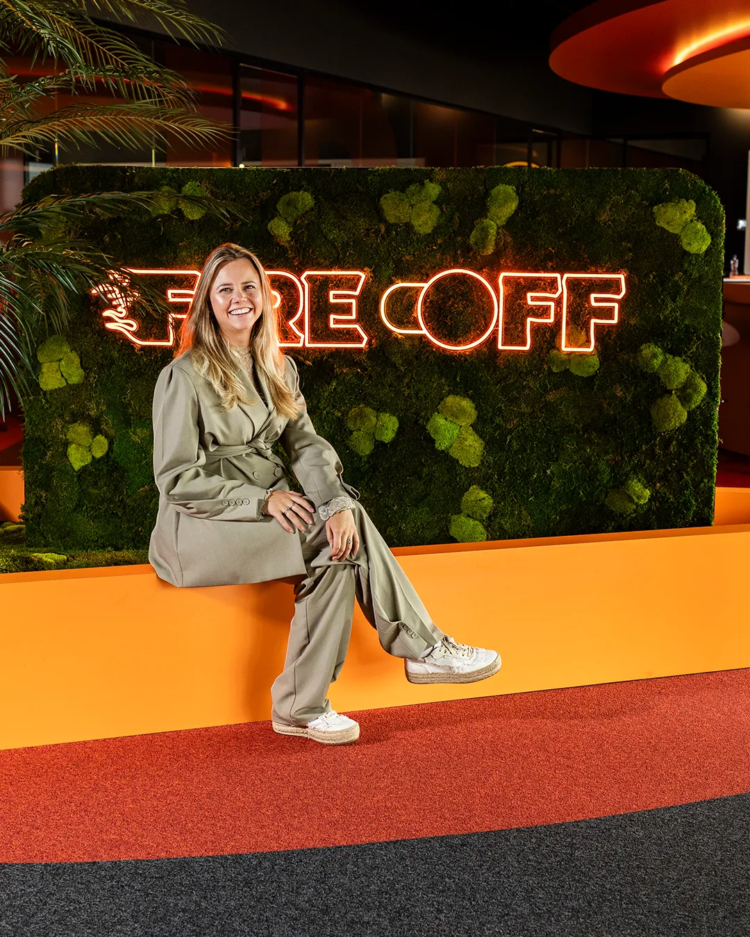 Professionele vrouw poseert bij FireOff neon logo met groene muur en oranje vloer tijdens bedrijfsevenement