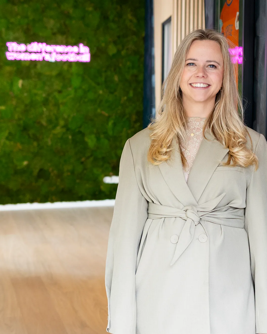 Blonde zakenvrouw in beige pak met brede glimlach voor groene muur met neon verlichting in modern kantoor