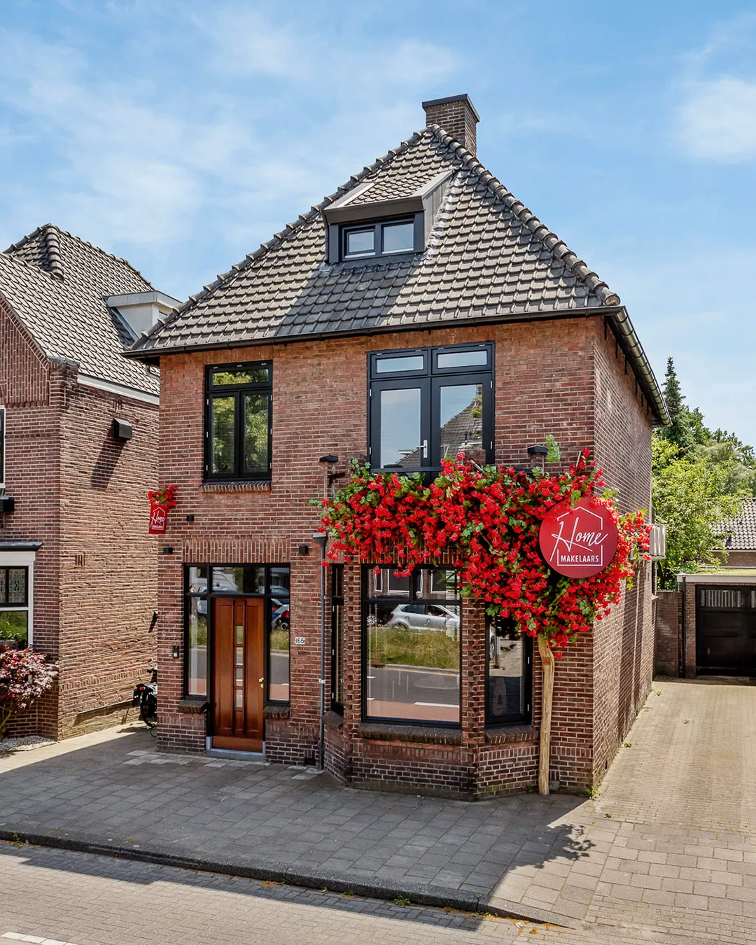 Karakteristiek bakstenen hoekpand met zadeldak en rode bloemendecoratie als makelaardijkantoor