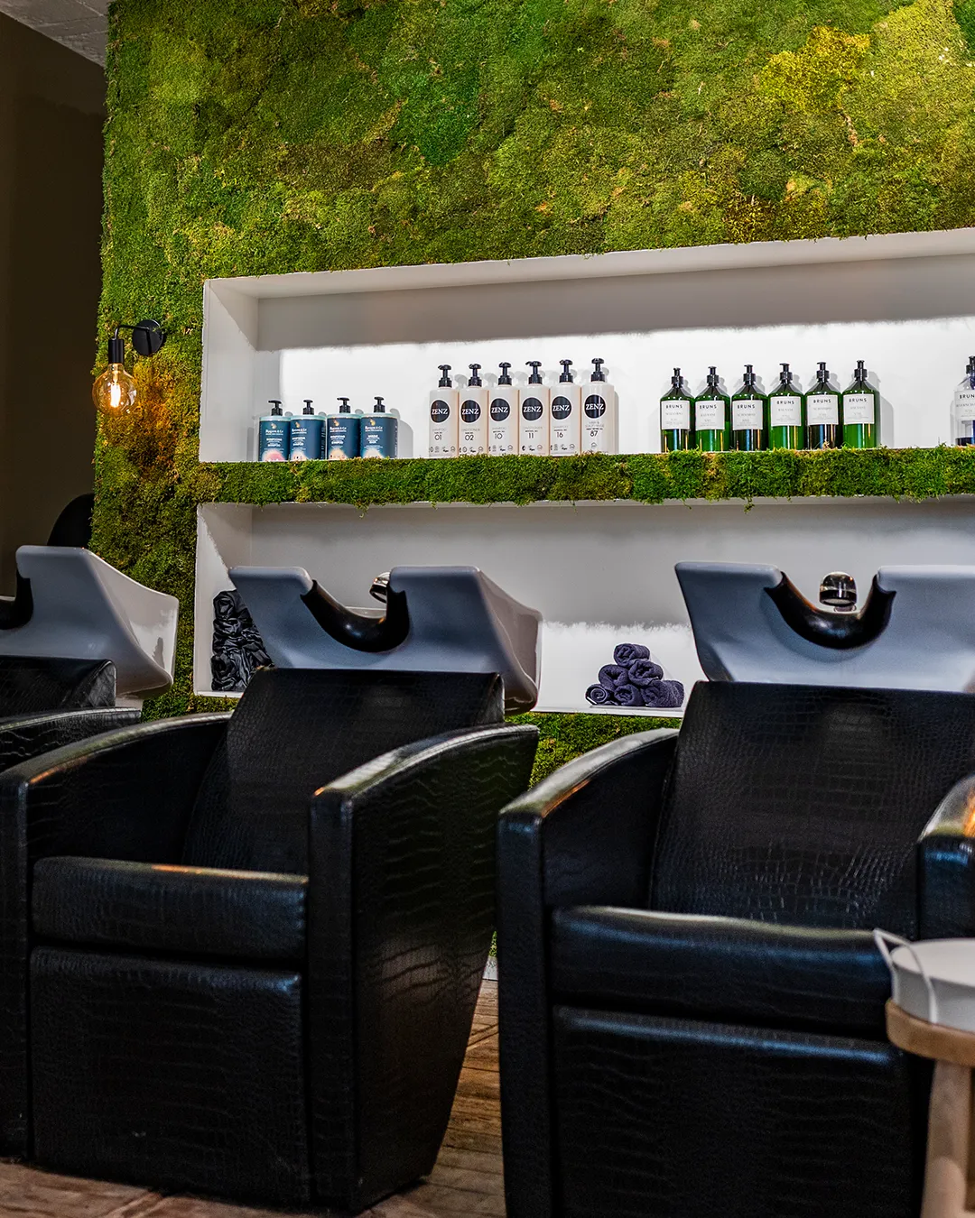 Moderne kapsalon met waszones, zwarte stoelen en verticale plantenwand met producten op witte planken