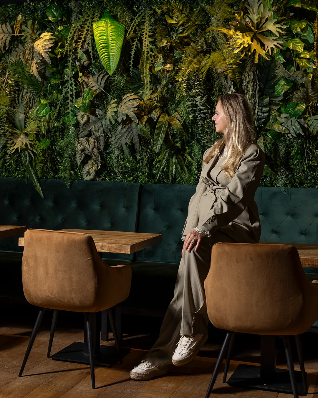 Vrouw in beige trenchcoat voor verticale plantenwand met tropische planten en luxe fluwelen banken