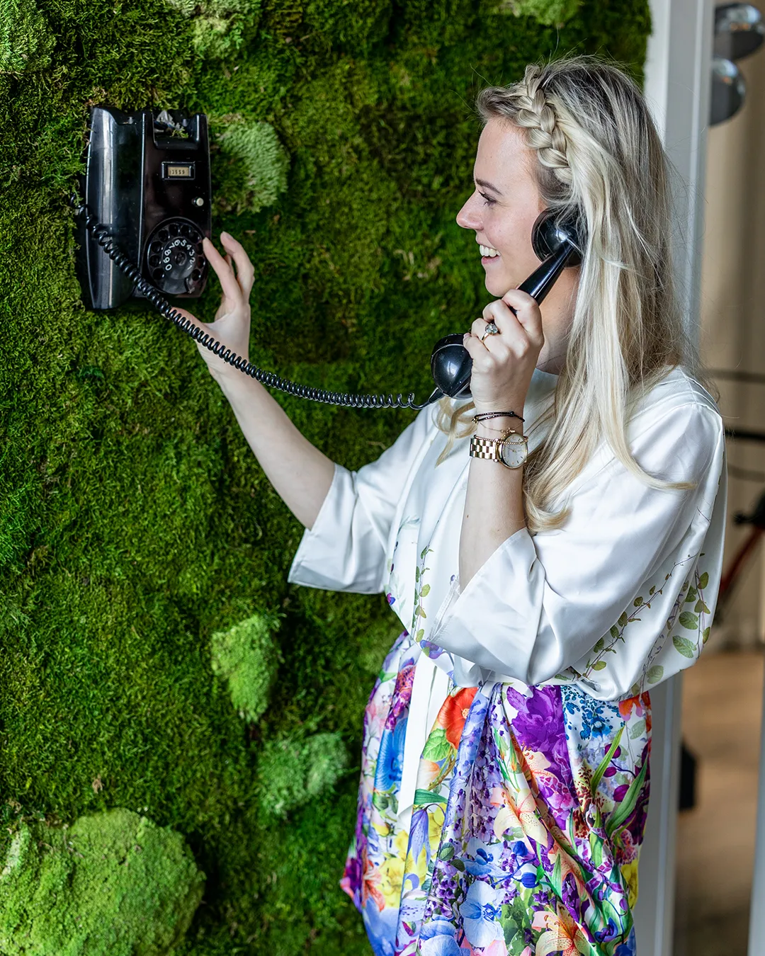 Blonde vrouw belt met zwarte vintage telefoon tegen groene plantenwand in modern interieur