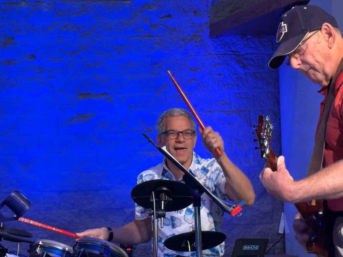 Smiling man playing electronic drums with red drumsticks and another man playing guitar wearing a cap against a blue-lit wall.