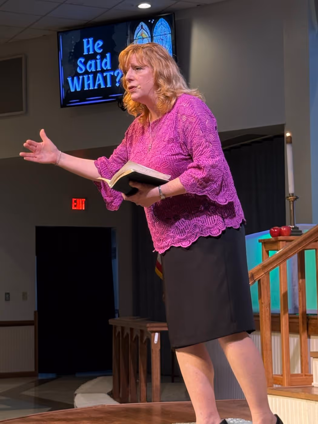 Woman in a purple blouse speaking passionately while holding an open book on a stage with a screen behind displaying the text 'He Said WHAT?'