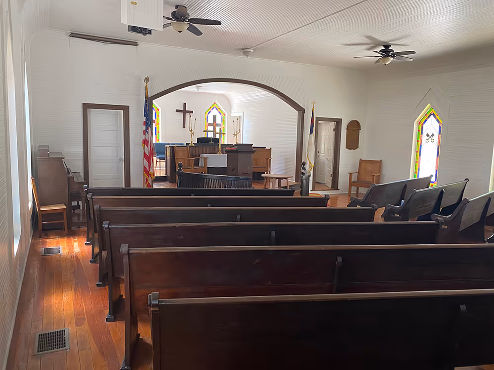 Interior of a small church with wooden pews, a piano on the left, stained glass windows, ceiling fans, an altar with a cross, American and Christian flags.