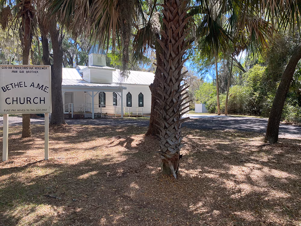 White Bethel A.M.E. Church building with a metal roof partially obscured by palm trees, and a sign in front stating the church was built in 1902 and moved to this site in 2002.