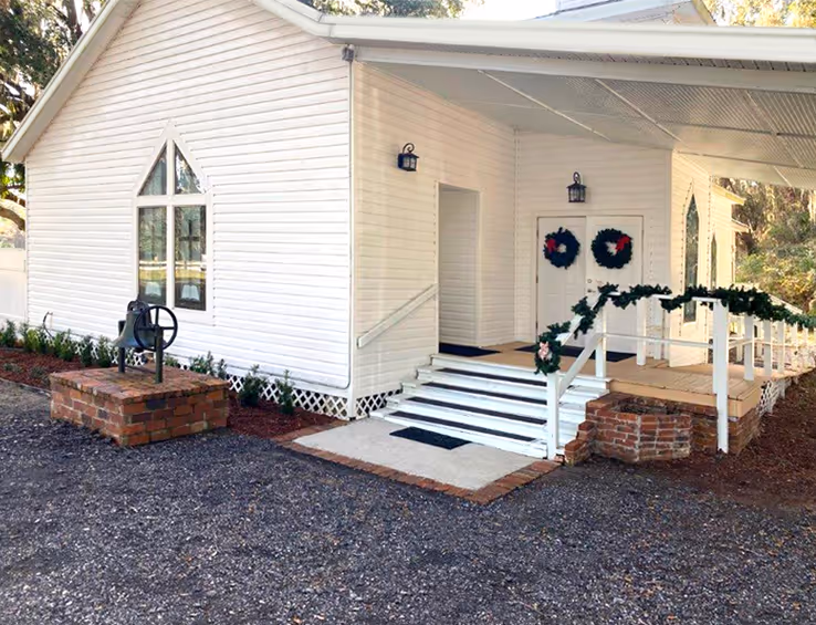 White wooden building with a covered porch decorated with Christmas wreaths and garlands, and a brick well next to it.
