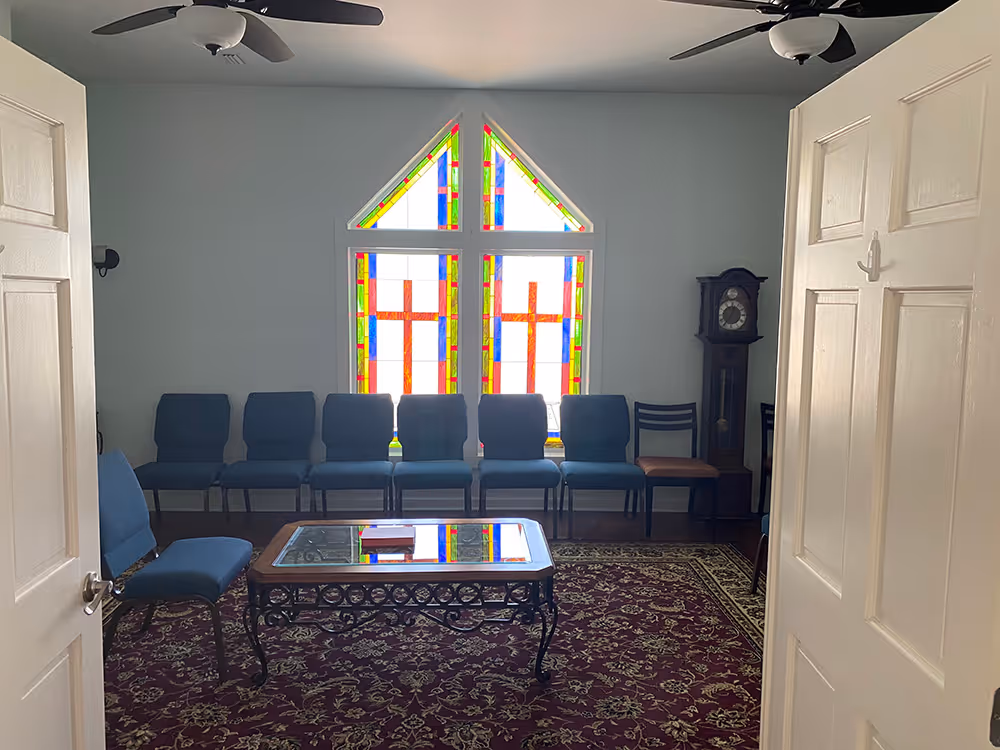 Room with open double doors showing blue chairs arranged in a row, a glass coffee table on a patterned red carpet, and a stained glass window with cross designs in the background.
