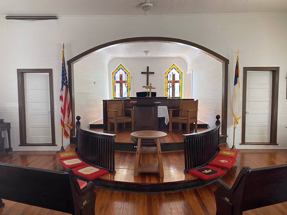 Interior view of a small church with wooden pews, pulpit, two stained glass windows with crosses, American and Christian flags, and red cushions with religious symbols on the floor.