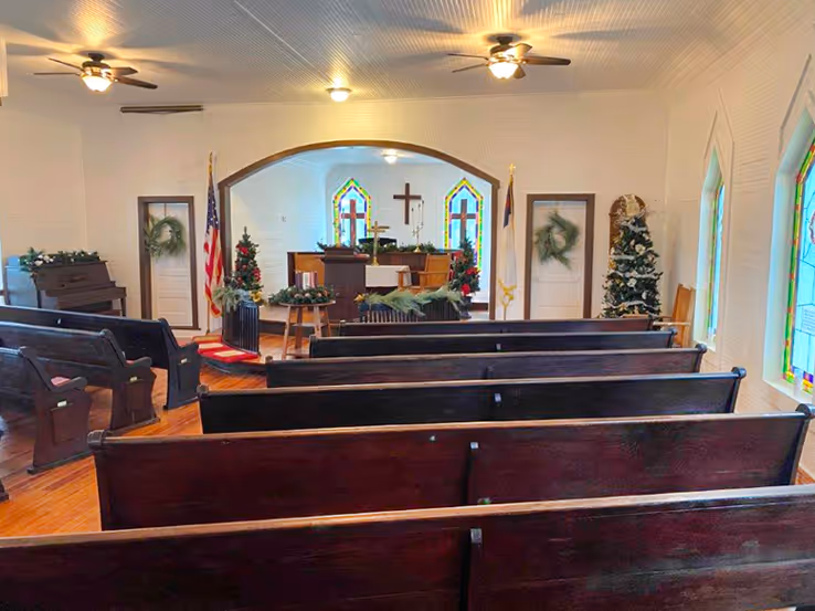Interior of a small church with wooden pews, stained glass windows, Christmas trees, and a cross above the altar area.