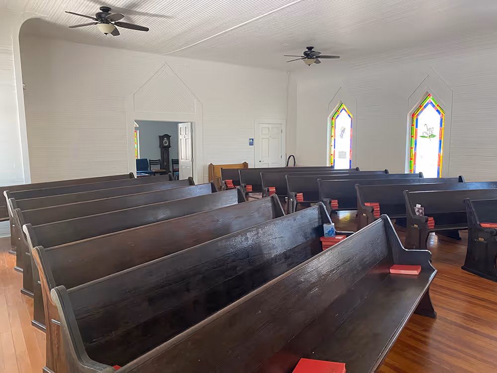 Empty church interior with wooden pews, red hymnals, stained glass windows, and ceiling fans.