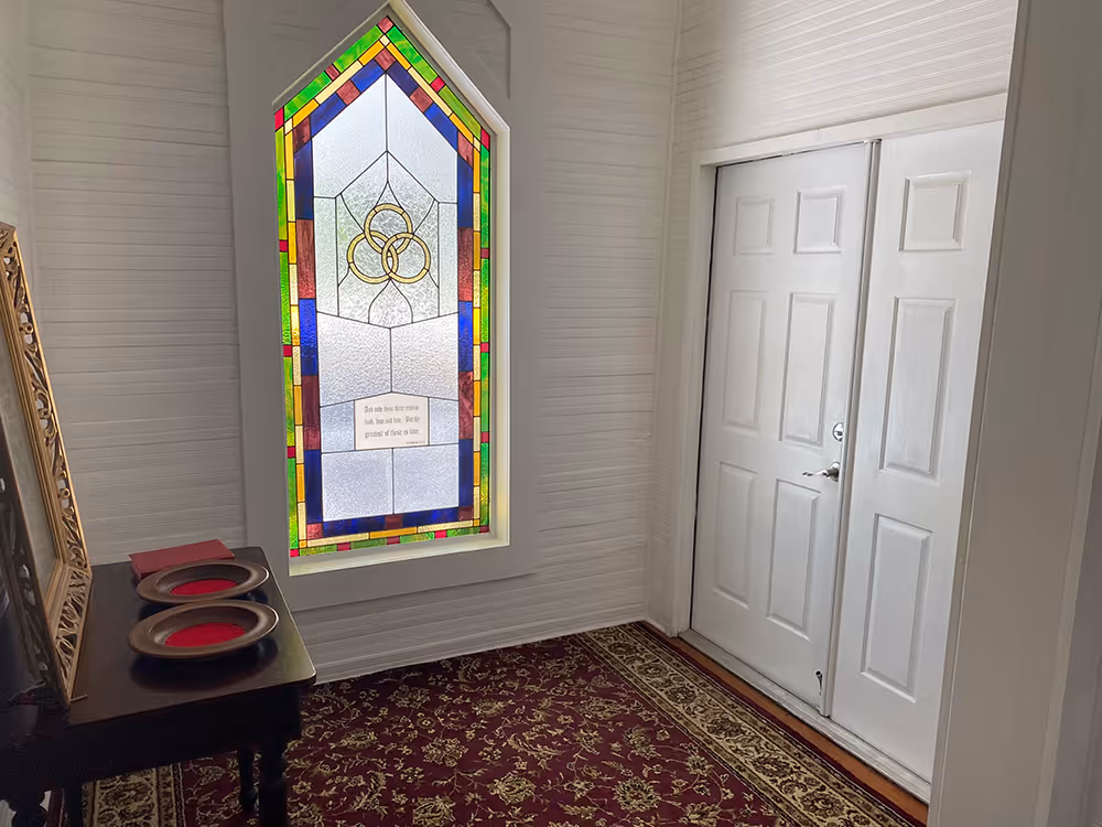Interior corner with a stained glass window featuring three interlocking rings and a Bible verse, a white double door, a dark wooden table with two offering plates, and a red and gold patterned carpet.