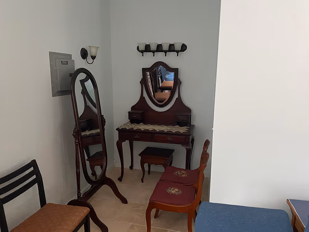 Corner of a room with vintage wooden vanity table with mirror, two floral cushioned chairs, a standing oval mirror, and a black chair with brown cushion.