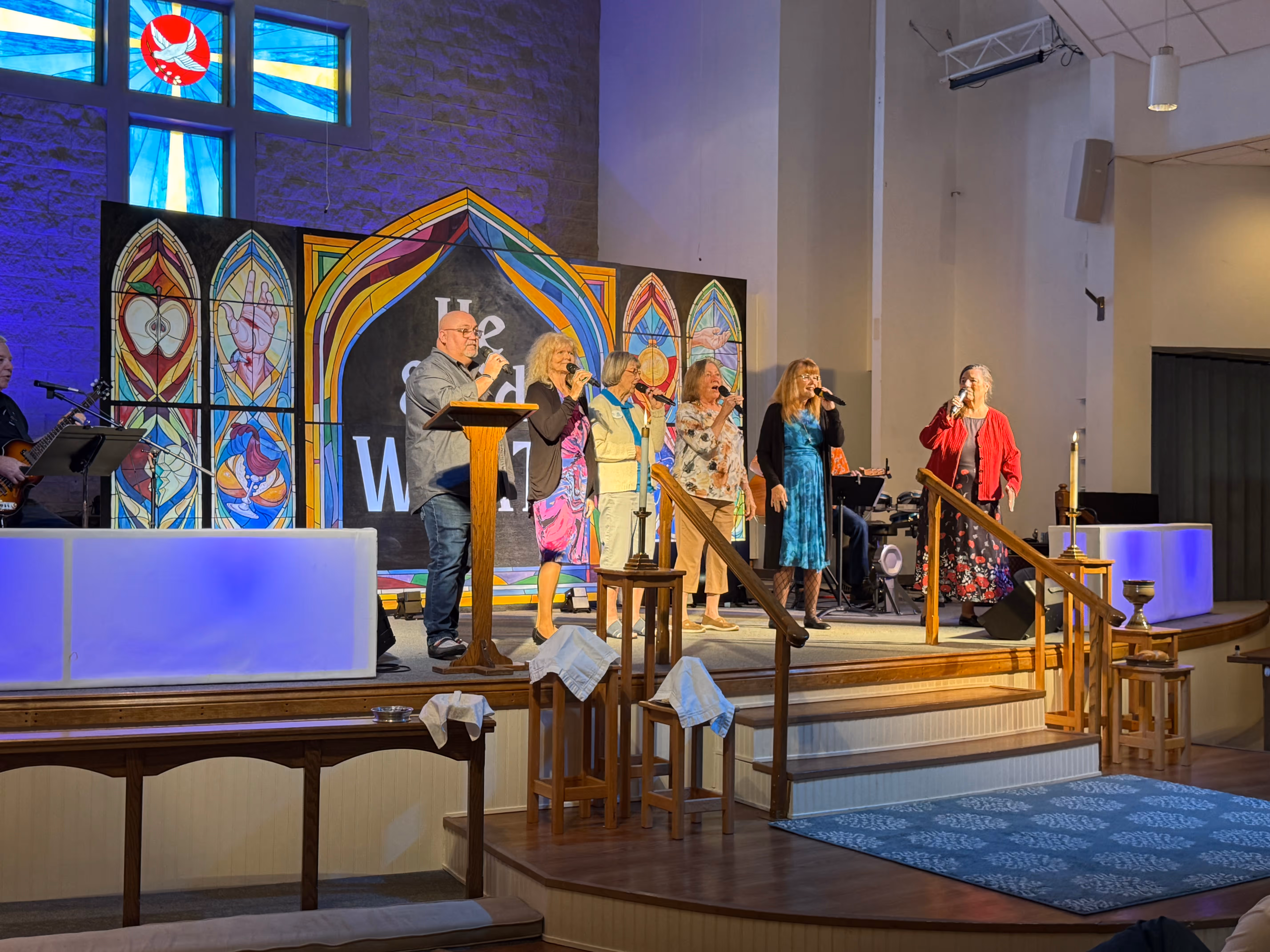 Group of six people singing with microphones on a church stage with colorful stained glass backdrop.
