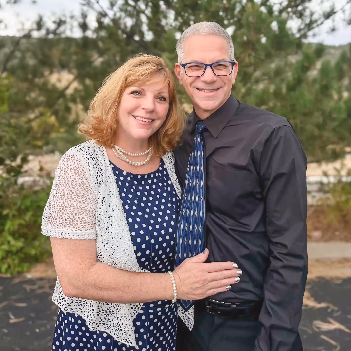 Smiling man in glasses wearing black shirt and patterned blue tie stands with woman in a blue polka dot dress and white lace shrug outdoors.