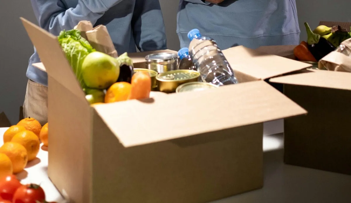 Cardboard box filled with assorted groceries including fresh vegetables, canned food, and bottled water on a table with people packing more boxes.