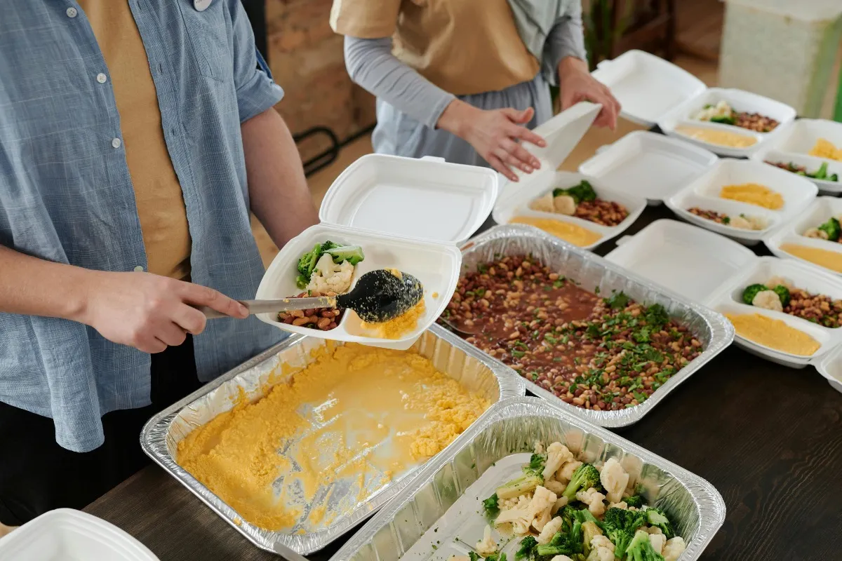 Two people serving food into white takeout containers including vegetables, beans, and a yellow dish from large aluminum trays.