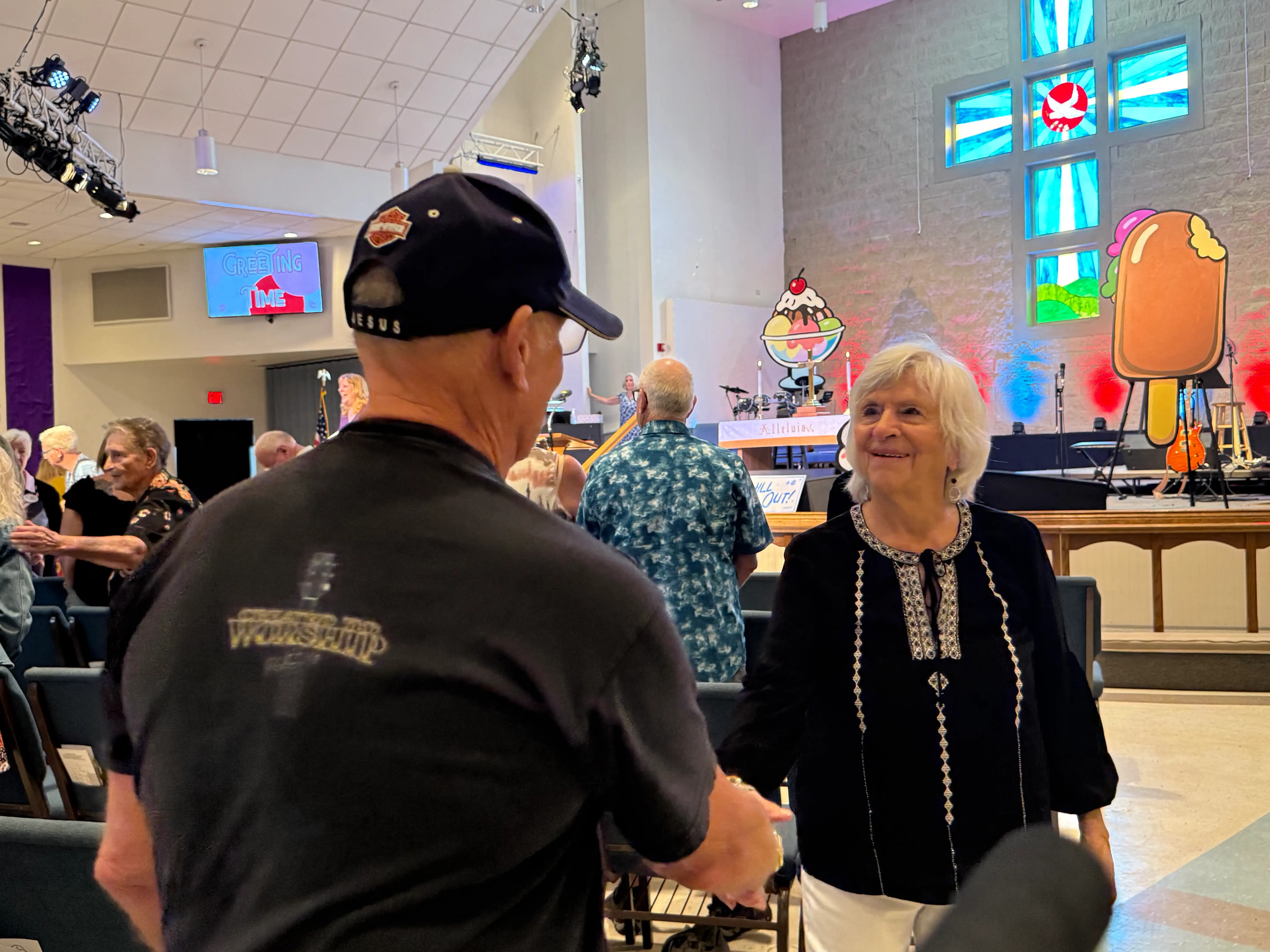 Two elderly people shaking hands and smiling inside a church with colorful stained glass windows and ice cream board decorations.