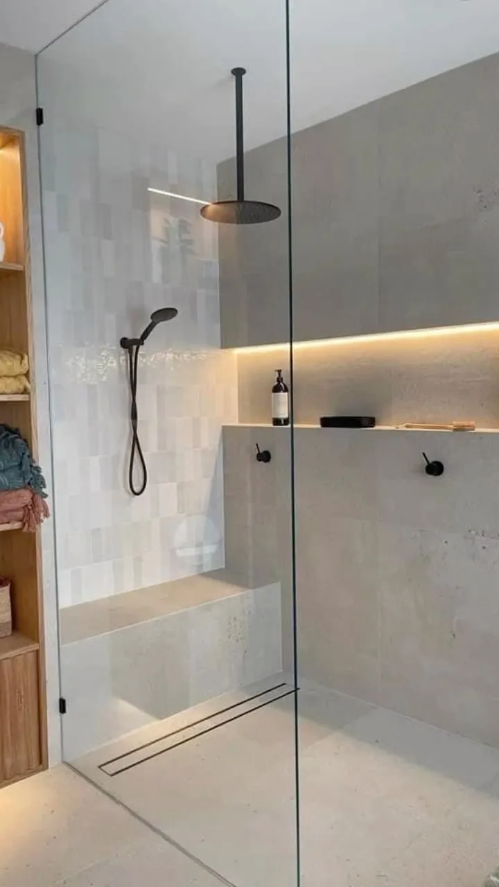 Minimalist walk-in shower with built-in bench, black rainfall and handheld showerheads, light gray tiles, and under-shelf LED lighting.