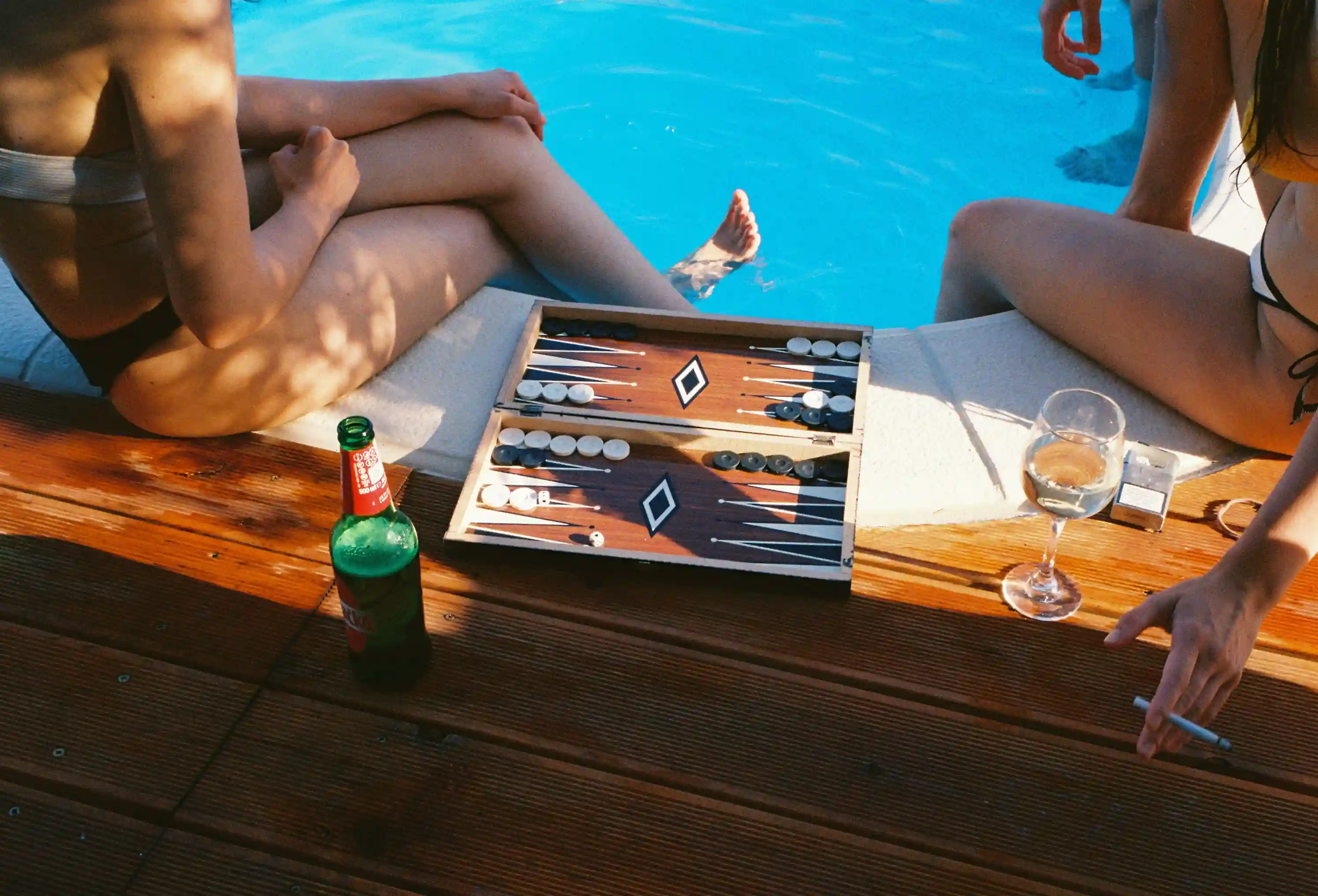 Two people sitting by a pool playing backgammon with a beer bottle and a wine glass nearby.