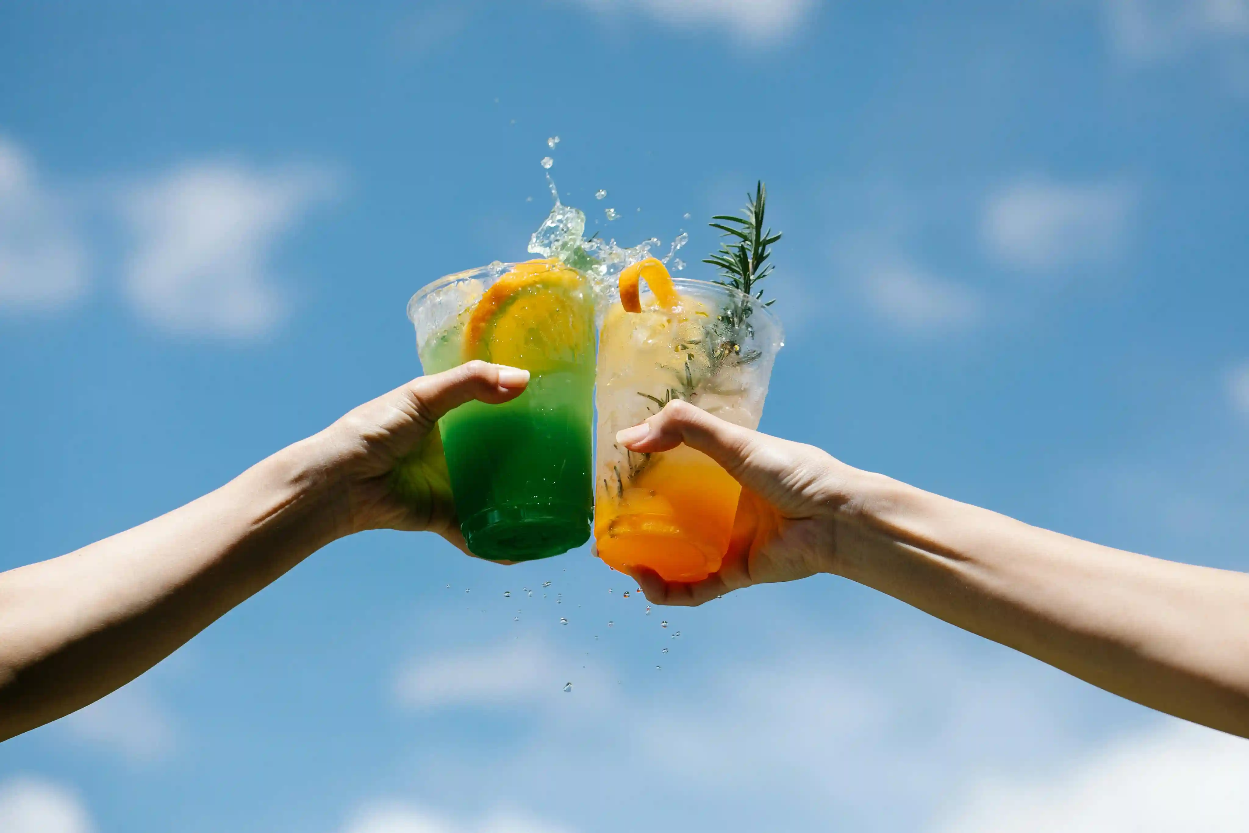 Two hands clinking plastic cups filled with colorful iced beverages against a blue sky.