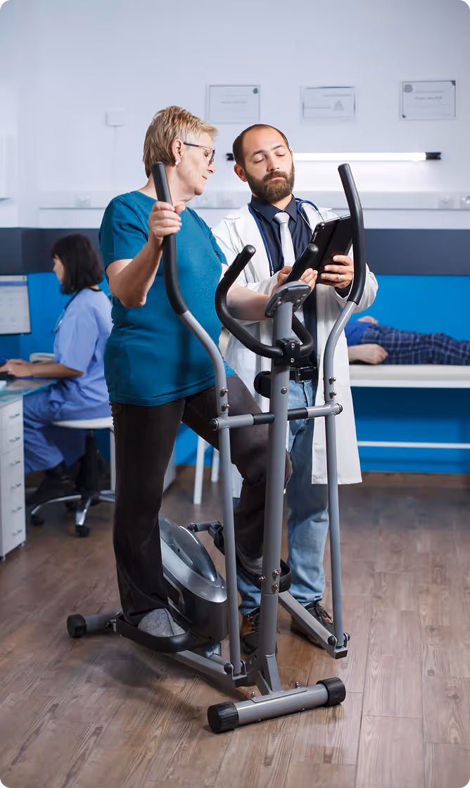 Senior woman exercising on an elliptical machine while a doctor reviews information on a tablet beside her in a clinical setting.