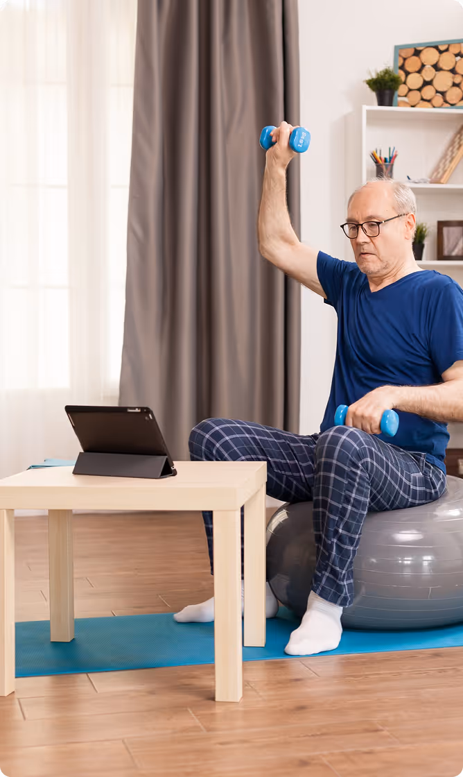 Elderly man wearing glasses and blue pajamas sitting on a gray exercise ball, lifting light blue dumbbells while following a tablet on a small wooden table.