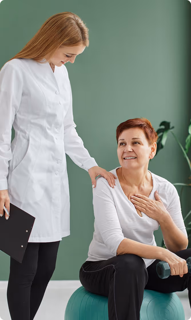 Healthcare professional in a white coat comforting a smiling older woman holding a dumbbell while sitting on an exercise ball.
