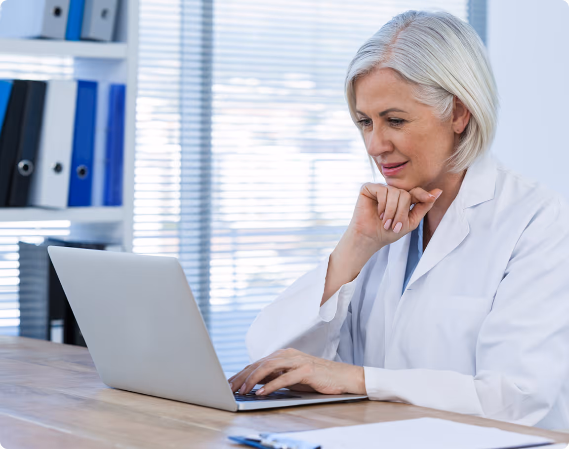 Mature female doctor with gray hair wearing a white coat, working on a laptop at a desk in an office.