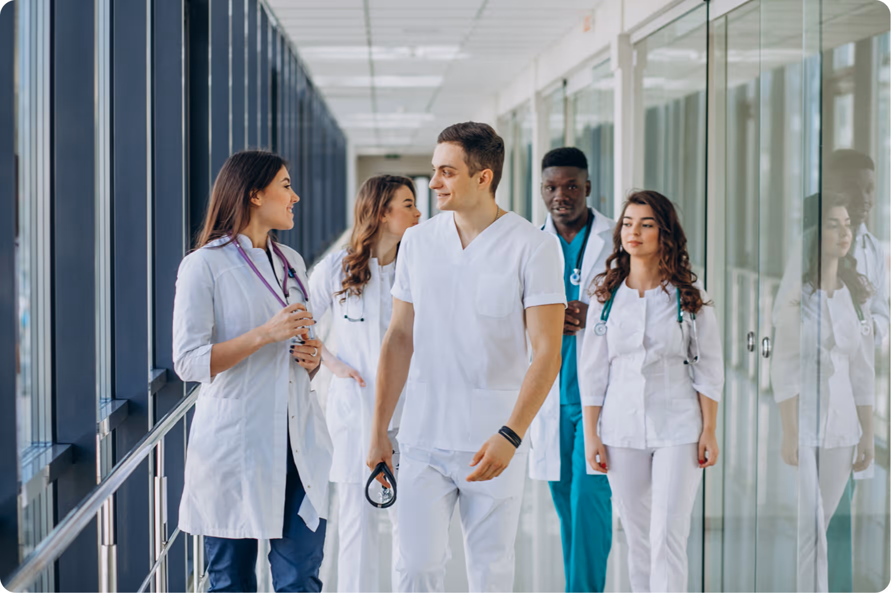 Five diverse doctors in white coats and scrubs walking and talking together in a hospital corridor.