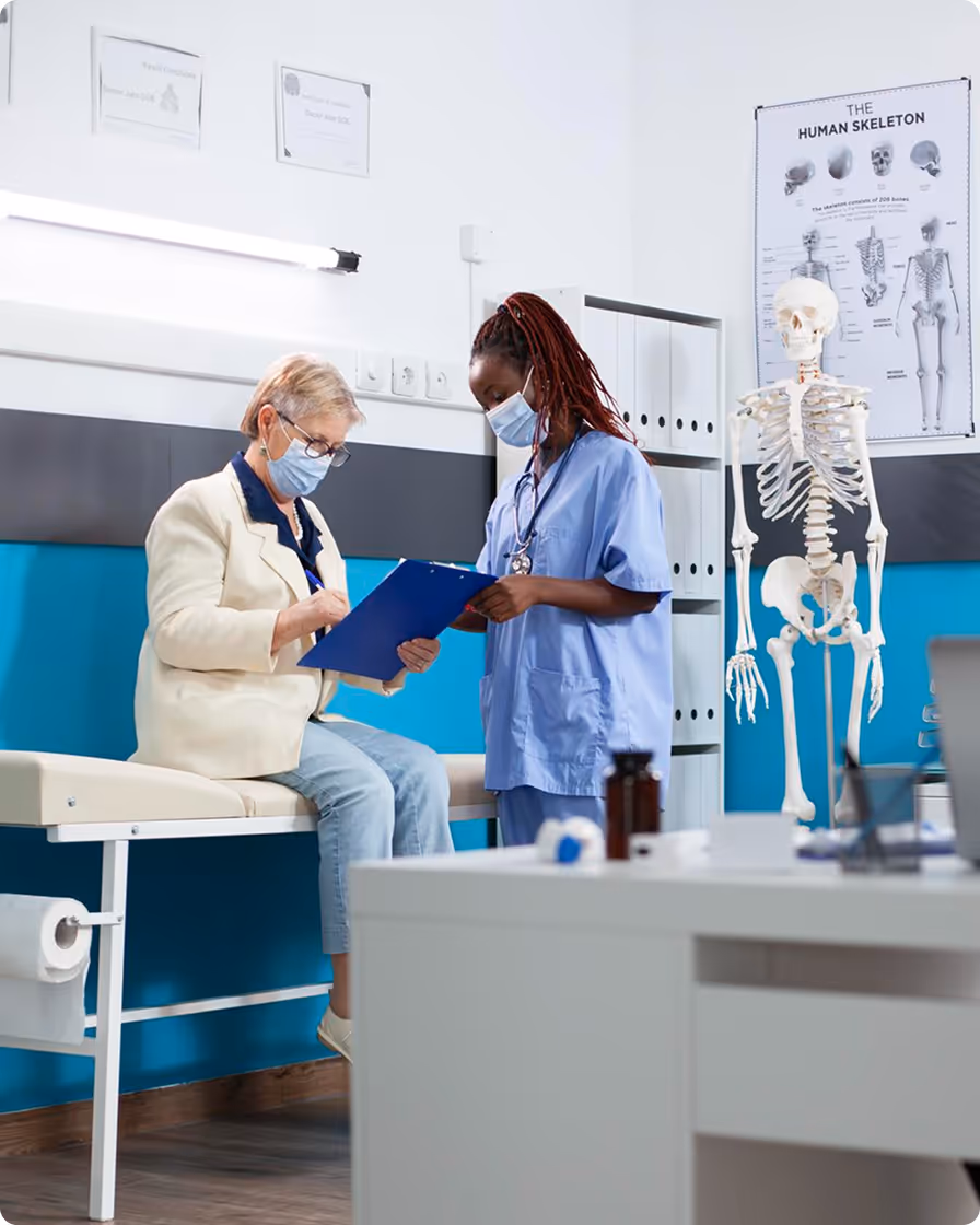 Medical professional in blue scrubs reviewing a clipboard with an elderly woman sitting on an exam table in a clinic room with a human skeleton model and anatomy poster in the background.
